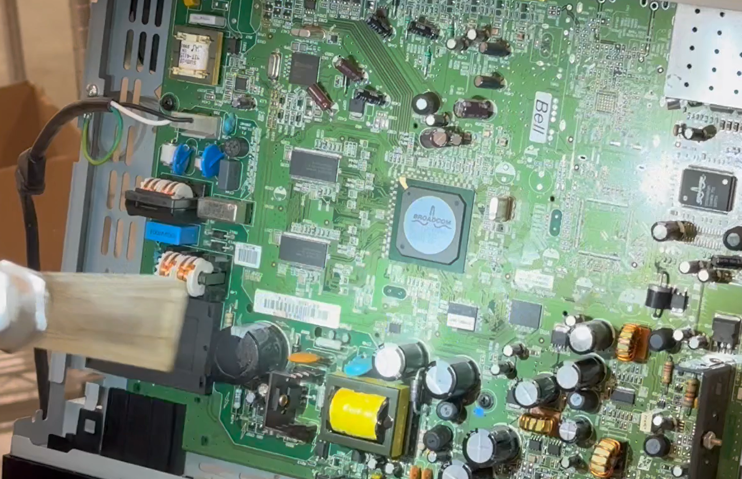 Eine grüne Leiterplatte mit verschiedenen elektronischen Bauteilen wie Kondensatoren, Chips und Steckverbindungen wird mit einer Trockeneisdüse gereinigt. Die Düse ist links im Bild zu sehen, während die feine Reinigung die Oberfläche der Leiterplatte von Staub und Schmutz befreit.