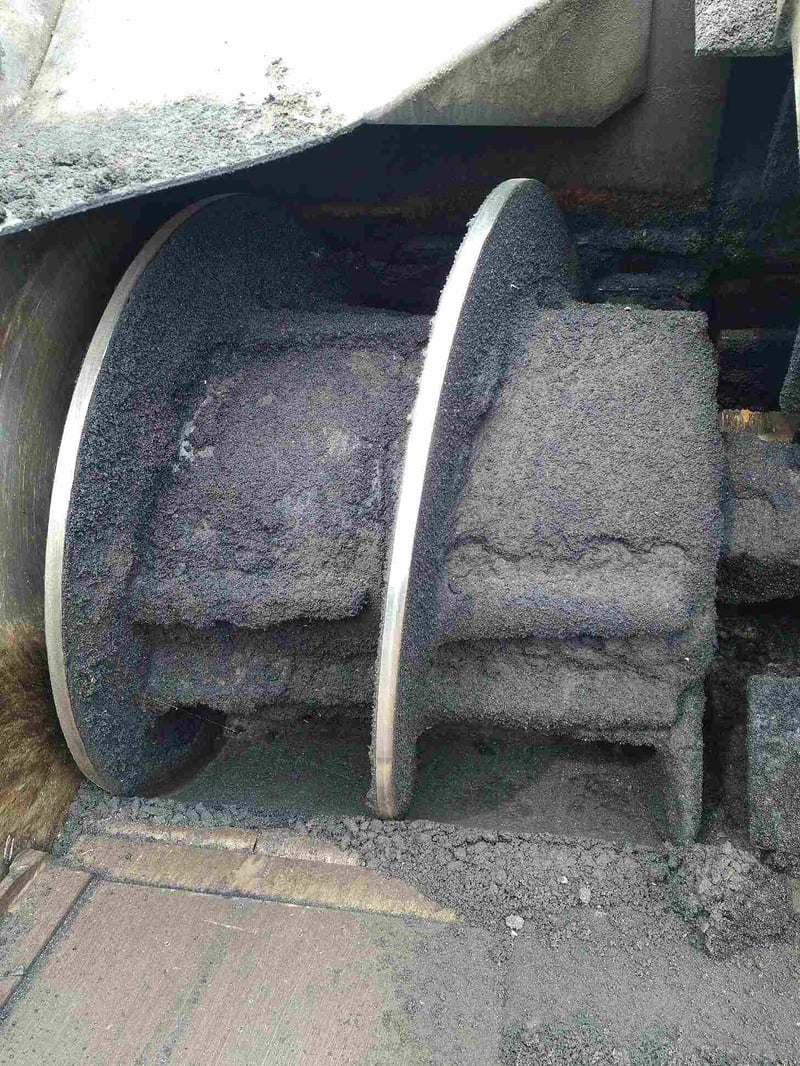 Nahaufnahme vor dem Trockeneisstrahlen einer großen, horizontalen Förderschnecke oder eines Verteilerelements einer Asphaltmaschine, das stark mit getrocknetem, grobem Asphaltmaterial bedeckt ist. Die Metalloberflächen der Schneckenflügel sind nur noch an den äußeren Kanten und den Verbindungsstücken sichtbar, der Rest ist von einer dicken, unregelmäßigen Schicht aus schwarzem, körnigem Asphalt verklebt. Am Boden der Maschine sammeln sich ebenfalls Asphaltreste.