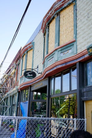 Außenansicht eines teilweise sanierten historischen Geschäftsgebäudes in Loveland, Ohio, nach einem Brand. Die oberen Fenster sind mit Holzplatten verschlossen, während im Erdgeschoss ein Restaurant mit dem Namen "Tano Bistro & Catering" geöffnet ist. Vor dem Gebäude sind Absperrungen und Gerüste zu sehen, was die Herausforderung der Brandschadensanierung mit Trockeneisstrahlen bei laufendem Betrieb verdeutlicht