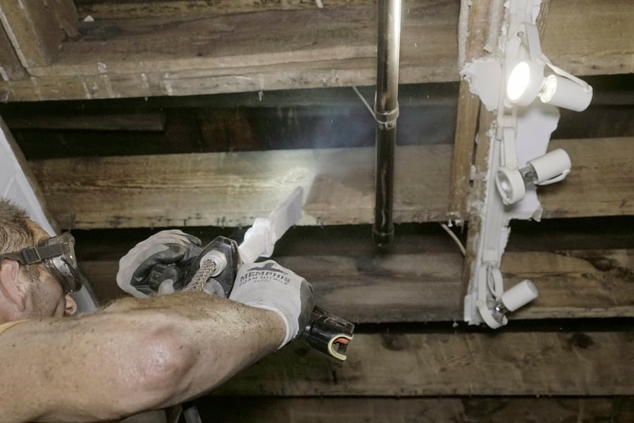 Ein Arbeiter mit Schutzbrille reinigt verkohlte Holzdeckenbalken in einem Gebäude nach einem Brandschaden. Er verwendet eine Trockeneisstrahlpistole, deren Strahl deutlich auf die Holzoberfläche trifft und den Ruß entfernt, ohne die Holzbalken zu beschädigen. Eine Metallleitung und Beleuchtungselemente sind ebenfalls an der Decke sichtbar.
