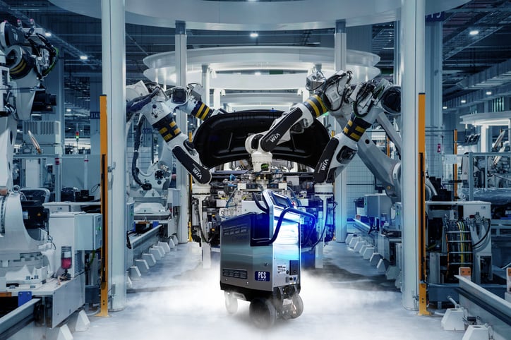 Moderne, hochautomatisierte Produktionslinie mit Robotern, die an einem Fahrzeug arbeiten. Im Vordergrund steht eine Trockeneisstrahlmaschine der PCS Ultra-Serie, umgeben von Nebel und blauem Licht. Symbolisiert innovative Reinigungstechnologie in der Automobilindustrie.