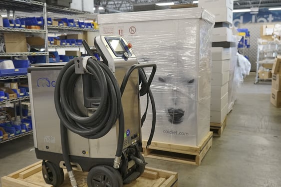 Eine Trockeneisstrahlmaschine vom Typ Cold Jet PCS 60 steht auf einer Holzpalette in einem Lagerraum. Im Hintergrund sind Regale mit blauen Behältern und verpackte Paletten mit dem Cold Jet-Logo zu sehen, was eine industrielle Umgebung darstellt.