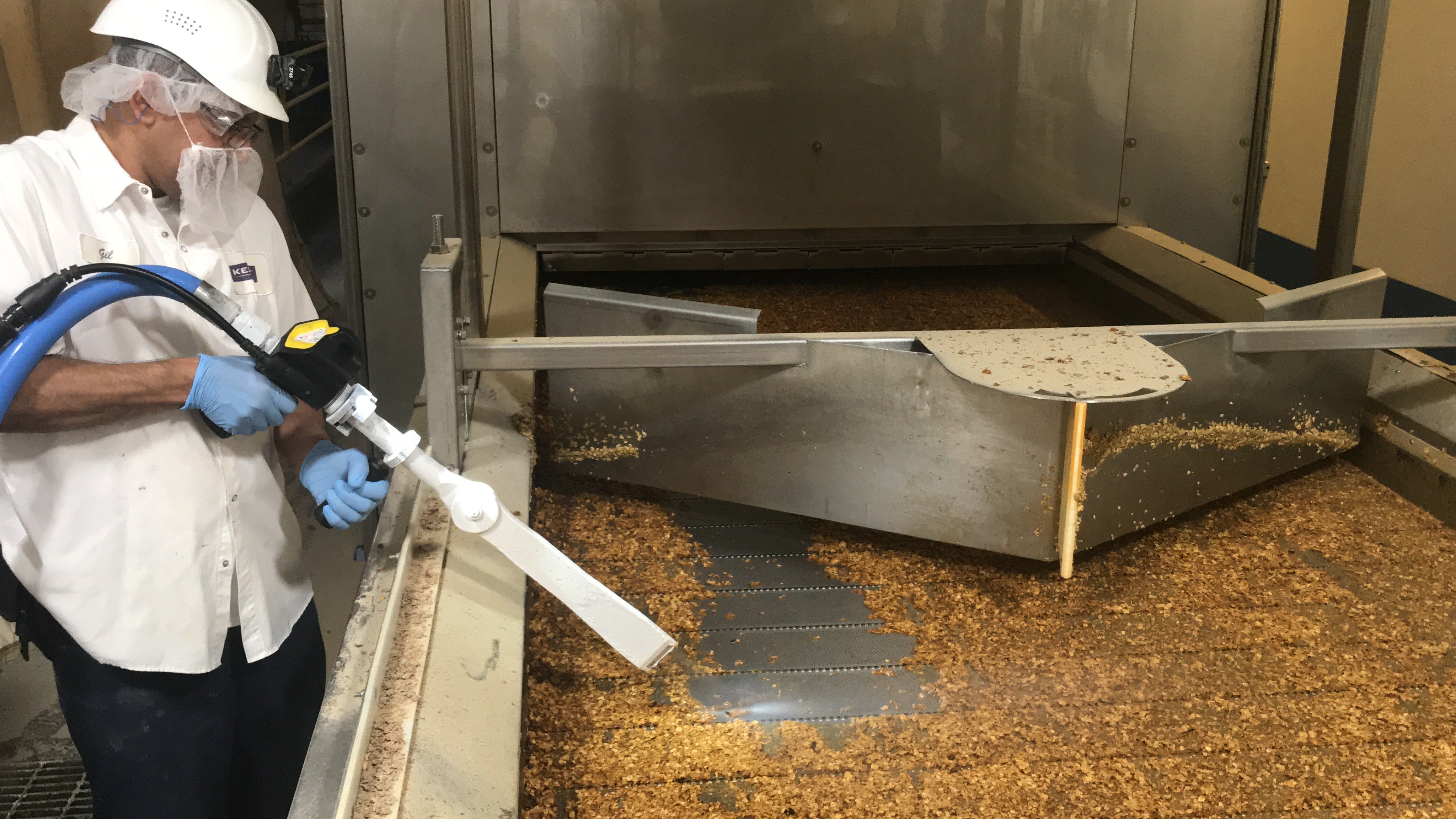 Ein Arbeiter in Schutzkleidung (weißes Hemd, Schutzhelm, Brille, Bartnetz und blaue Handschuhe) verwendet ein Trockeneisstrahlgerät mit einer breiten Düse, um eine große industrielle Reinigungsmaschine oder einen Förderer zu reinigen. Die Oberfläche des Förderers ist mit einer dicken Schicht aus braunen, körnigen Rückständen (vermutlich Lebensmittelreste) bedeckt.