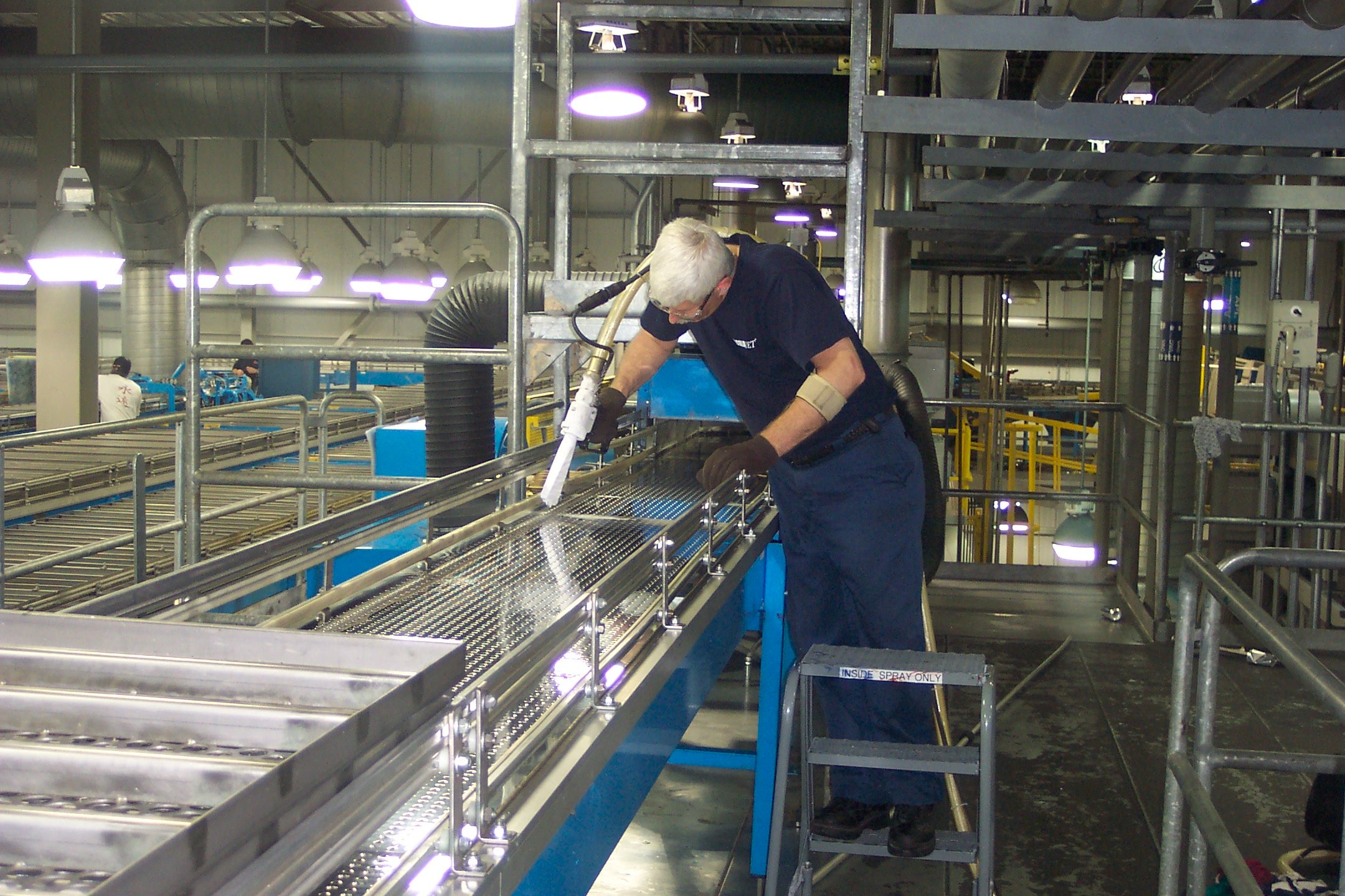 Ein Mitarbeiter reinigt mit einem Trockeneisstrahlgerät ein Förderband in einer industriellen Großbäckerei. Die Reinigung erfolgt ohne Wasser, direkt in der Produktionsumgebung
